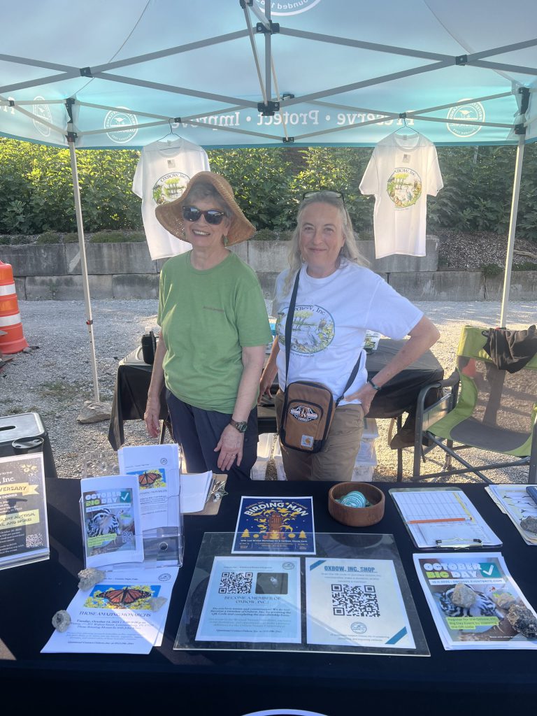 Two Oxbow volunteers smiling behind a table of brochures under a tent