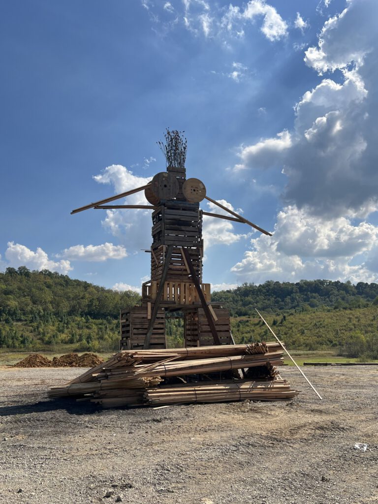 The wooden Birding Man effigy standing on a gravel lot under a blue sky